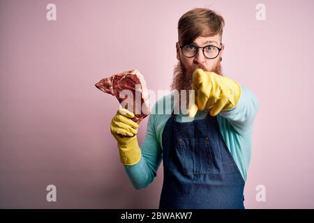 Rotkopf Irish Butcher man mit Bart, der rohes Rindersteak über rosafarbenem Hintergrund hält und mit dem Finger auf die Kamera und auf Sie zeigt, Handzeichen, positiv an Stockfoto