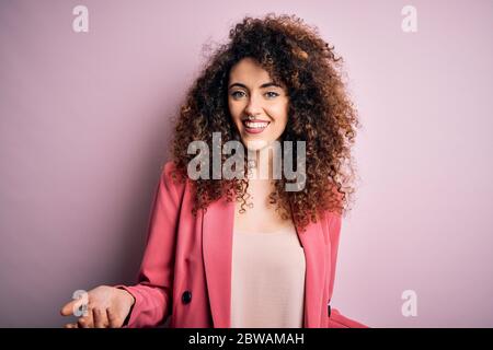 Junge schöne Geschäftsfrau mit lockigen Haaren und Piercing tragen elegante Jacke lächelnd fröhlich mit offenen Armen als freundliche Begrüßung, positiv und Co Stockfoto