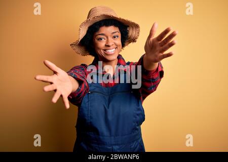 Junge afro amerikanische Afro Farmer Frau mit lockigen Haaren trägt Schürze und Hut Blick auf die Kamera lächelnd mit offenen Armen für Umarmung. Fröhliche Expressi Stockfoto