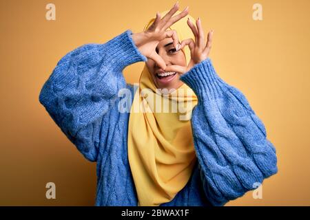 Junge schöne brünette muslimische Frau trägt arabischen Hijab über isoliert gelben Hintergrund tun Herzform mit Hand und Finger lächelnd suchen throu Stockfoto