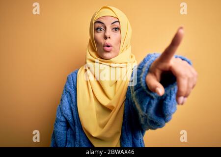Junge schöne Brünette muslimische Frau trägt arabischen Hijab über isoliert gelben Hintergrund zeigt mit Finger überrascht voraus, offenen Mund erstaunt expre Stockfoto