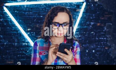 Porträt der charmanten Frau durch verschiedene Nachrichten im Handy gegen beleuchtete Wand blättern. Stockfoto