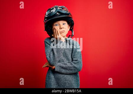 Junge kleine kaukasische Kind trägt Vintage Biker Motorradhelm und googles über rotem Hintergrund denken, müde und gelangweilt mit Depression PR Stockfoto