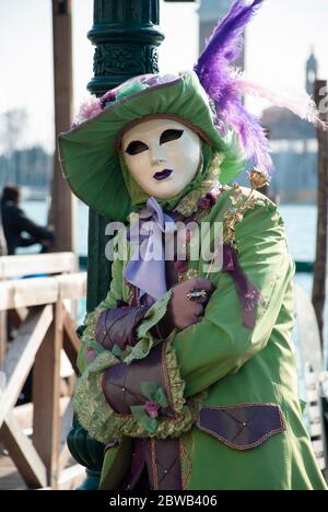 Eine Person, die sich während des Karnevals in Venedig in Italien an einem Lampenkosten am großen Kanal in grüner Verkleidung und Maske lehnt. Stockfoto