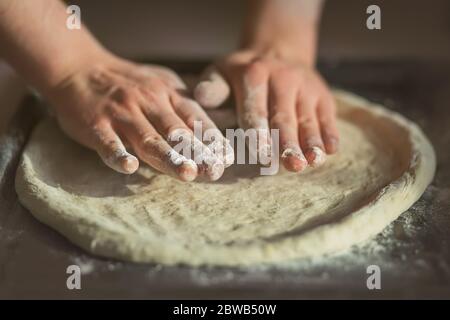 Der Koch rollt den Teig für hausgemachte leckere Pizza auf einem dunklen Backblech aus, mit Mehl bestreut. Hausmannskost. Stockfoto