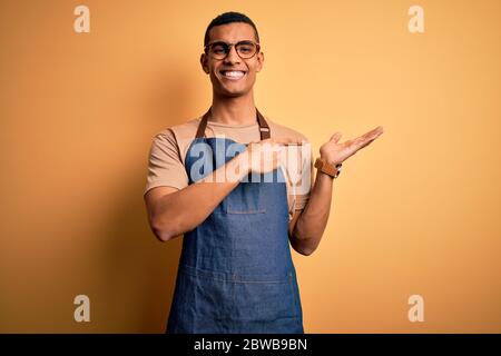 Junge gut aussehende afroamerikanischen Ladenbesitzer Mann trägt Schürze über gelbem Hintergrund erstaunt und lächelnd an die Kamera, während mit Hand und po präsentieren Stockfoto
