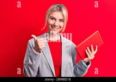 Junge schöne blonde Geschäftsfrau arbeitet mit Tablet über isolierten roten Hintergrund lächelnd glücklich und positiv, Daumen nach oben tun ausgezeichnet und approv Stockfoto