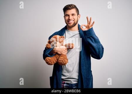 Junge blonde Mann mit Bart und blauen Augen tragen Pyjama halten Teddybär tun ok Zeichen mit Fingern, ausgezeichnetes Symbol Stockfoto