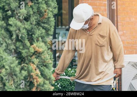 Ein alter Mann mit Quarantäne trägt eine Gesichtsmaske, um die Ausbreitung des Corona-Virus (Covid-19) zu verhindern, das Pflanzen für die Bewegung im Garten zu Hause bewässert. Nein Stockfoto