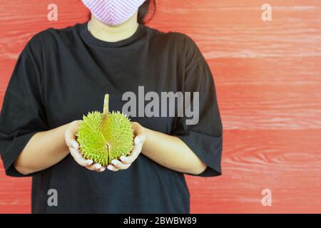 Junge Frau trägt eine Schutzmaske, um eine Infektion des Corona-Virus (Covid-19) in einem schwarzen Hemd mit einem reifen Durian zu verhindern, bereit, mit dem roten w zu essen Stockfoto