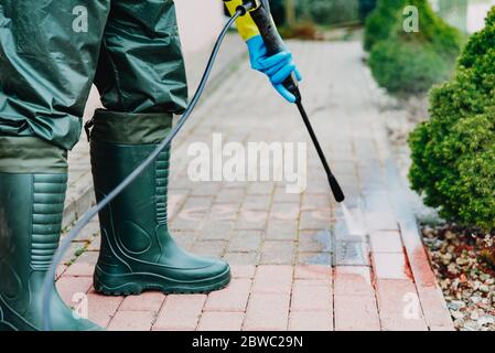 Mann putzt roten, conrete Bürgersteig Block mit Hochdruck-Wasserdeck. Reinigungskonzept für Pflaster. Mann mit Wathosen, wasserdichte Schutzhose Stockfoto