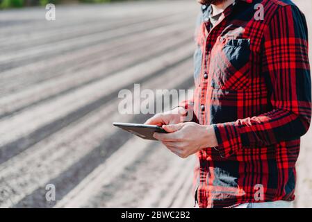 Farmer in rot kariertem Hemd mit Tablet auf trockenem Ackerland. Anwendung moderner Technologien und Anwendungen in der Landwirtschaft. Konzept der intelligenten Landwirtschaft und agr Stockfoto