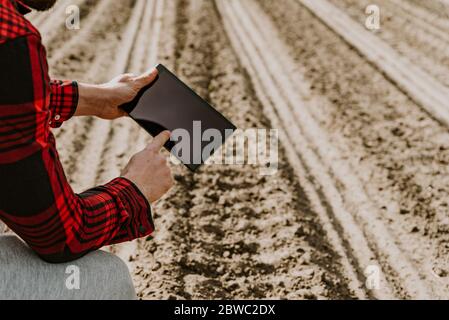 Farmer in rot kariertem Hemd mit Tablet auf trockenem Ackerland. Anwendung moderner Technologien und Anwendungen in der Landwirtschaft. Konzept der intelligenten Landwirtschaft und agr Stockfoto