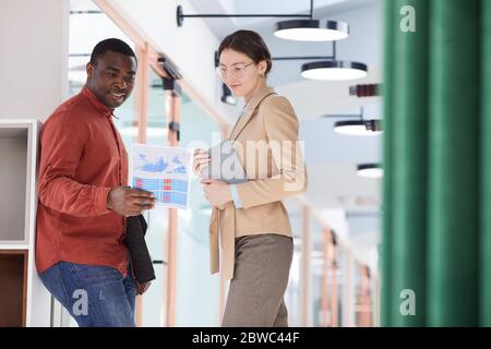 Porträt eines lächelnden afroamerikanischen Mannes, der Dokumente hält und mit einer eleganten jungen Geschäftsfrau im zeitgenössischen Bürogebäude spricht Stockfoto