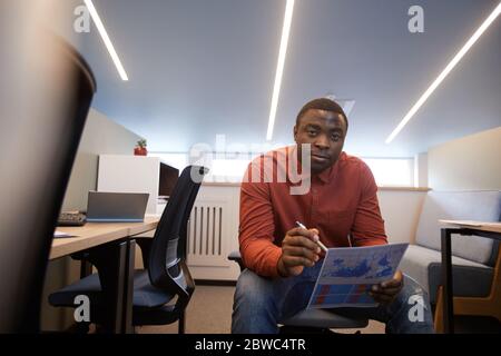 Weitwinkelporträt des zeitgenössischen afroamerikanischen Mannes, der ein Dokument hält und die Kamera betrachtet, während er auf einem Stuhl in einem futuristischen Bürogebäude sitzt, Stockfoto