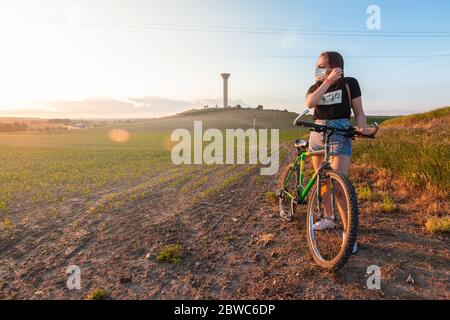 Junge kaukasische Frau auf einem Fahrrad mit chirurgischer Maske in einer schönen Landschaft mit grünen Feldern zum Horizont und blauen Himmel mit Wolken. Stockfoto