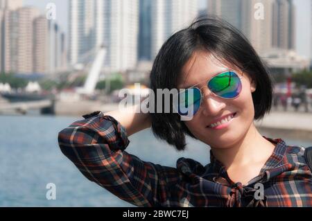 Eine lächelnde chinesin, die ihre Haare in einer Sonnenbrille hält und die Stadt Qingdao China im Hintergrund verschwommen. Stockfoto