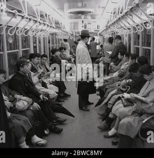 1960er, historisch, Japan, Tokyo Metro, japanische Pendler, die in einer Kutsche mit einer U-Bahn oder U-Bahn fahren, die meisten sitzen, einige mit Regenschirmen, da starke Regenstürme ein häufigen Ereignis in der Stadt sind. Stockfoto