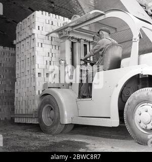 1950s, historischer, männlicher Fahrer, der am Steuer eines Gabelstaplers der Ära sitzt, der eine schwere Ladung von neu produzierten Londoner Ziegelsteinen bewegt, Stewartby, Bedfordshire, England, Großbritannien. Batteriebetriebene Gabelstapler waren offene Cockpit-Flurförderzeuge, die verwendet wurden, um Materialien kurze Wege zu bewegen. Stockfoto