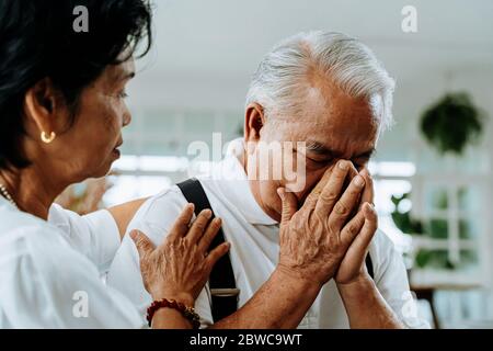 Unglücklich im Ruhestand asiatische ältere Paar sitzt auf dem Sofa, während ältere Frau tröstlich ihren traurigen Mann zu Hause Stockfoto