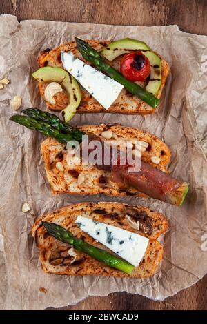 Hausgemachtes geröstetes Brot mit grünem Spargel, eingewickelt in Speck, Käse, Avocado-Scheiben, gegrillte Tomatenkirsche und Nüsse. Draufsicht Stockfoto