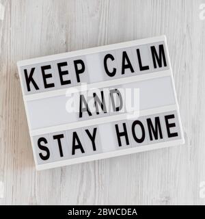 „Keep Calm and Stay Home“-Worte auf einem modernen Brett auf weißem Holzhintergrund, Draufsicht. Über Kopf, von oben, flach gelegt. Stockfoto