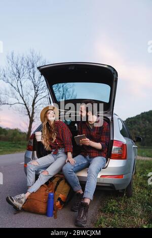 Paar sitzen auf dem Kofferraum des Autos und staunen über Foto auf Tablet. Mädchen und Jungen reden, lächeln und Spaß. Time to Travel Konzept Stockfoto