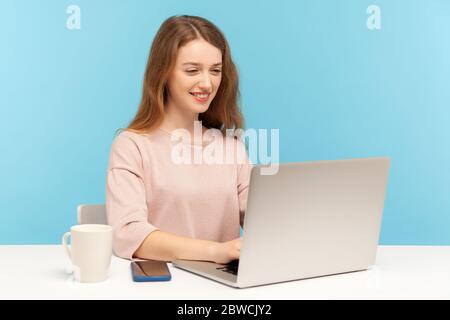 Fröhliche junge Geschäftsfrau in Freizeitkleidung sitzt am Arbeitsplatz, tippen auf Laptop und lächeln, online arbeiten, tun freiberufliche Arbeit im Home Office. Stockfoto