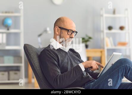Ein kluger, reifer Mann mit Brille sieht in einem Tablet in seinen Händen, während er in einem bequemen Sessel im Wohnzimmer sitzt. Stockfoto