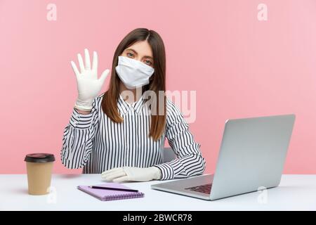 Freundliche Geschäftsfrau sitzt sicher gesund mit hygienischen Maske und Schutzhandschuhe während der Quarantäne, winken Hallo Geste. Arbeiten im Heimbüro i Stockfoto