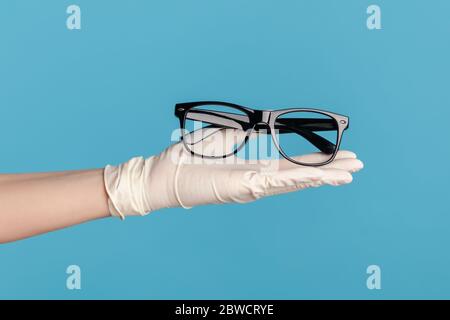 Profil Seitenansicht Nahaufnahme der menschlichen Hand in weißen chirurgischen Handschuhen halten und geben schwarze Brillen Rahmen. Indoor, Studio-Aufnahme, isoliert auf blauem Rücken Stockfoto