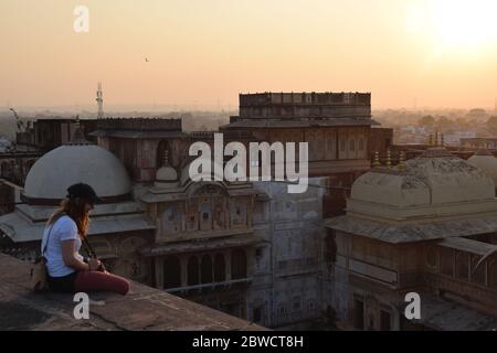 Junge Frau und Touristen, die auf dem Dach sitzt und den Sonnenuntergang über der Skyline der Stadt mit einer Kamera um den Hals beobachtet. Foto aufgenommen in Indien. Stockfoto