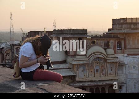 Alleinreisende und junge Frau, die ihre Kamera-Ausrüstung repariert, auf dem Dach sitzend. Wunderschöner Sonnenuntergang und Skyline der Stadt. Foto aufgenommen in Indien. Stockfoto