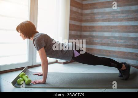 Erwachsene fit schlanke Frau hat Training zu Hause. Seitenansicht des starken, gut gebauten Senior-Modellständers in voller Plankenposition. Stretching Rücken und Körper. Stockfoto