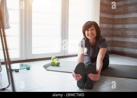 Erwachsene fit schlanke Frau hat Training zu Hause. Fröhliche positive reife Person, die den Oberkörper nach vorne in Richtung Zehen streckt und lächelt. Posing auf der Kamera. Nehmen Stockfoto