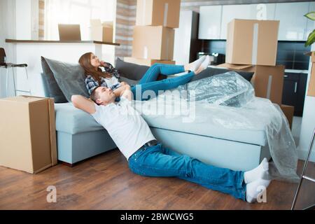 Pärchen auf Couch nach dem Einzug, Mann und Frau auf Sofa entspannen gerade in Wohnung mit Pappkartons auf dem Boden gezogen, glücklich zufrieden Stockfoto