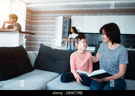 Großmutter liest ihrer Enkelin ein Buch vor. Vater arbeitet an einem Computer. Mutter kocht in der Küche. Große Familie zu Hause. Stockfoto