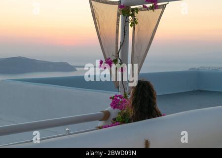 Frau sitzt auf einer Bank unter einer Pergola mit einer Voukamvilia Pflanze und genießt den atemberaubenden Blick auf den Sonnenuntergang auf Santorini Stockfoto