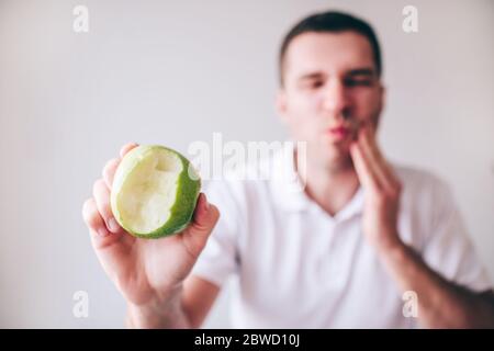 Junger Mann in weißem Hemd isoliert über verschwommenem unscharf Hintergrund. Guy leiden unter Schmerzen Zahnschmerzen und Schmerzen. Zeigen Sie gebissenen grünen Apfel auf der Kamera. Stockfoto