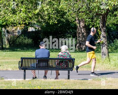 Ein altes Paar sitzt auf einer Parkbank und ein Mann mittleren Alters läuft mit Kopfhörern im Park Stockfoto