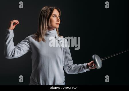 Schöne Schwertfrau im Fechtanzug hält Rapier und Blick weg isoliert auf schwarz Stockfoto