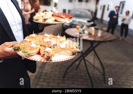 Mann in formeller Kleidung trägt Teller mit verschiedenen Mini-Sandwiches. Kleine leckere Snacks. Feier, Party, Geburtstag oder Hochzeit Konzept. Stockfoto