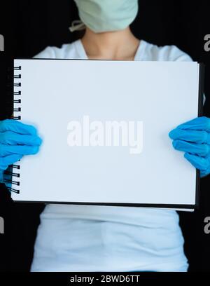 Nahaufnahme einer leeren Notizbuchseite, die von einer Frau mit Gesichtsmaske und OP-Handschuhen mit schwarzem Hintergrund gehalten wird Stockfoto