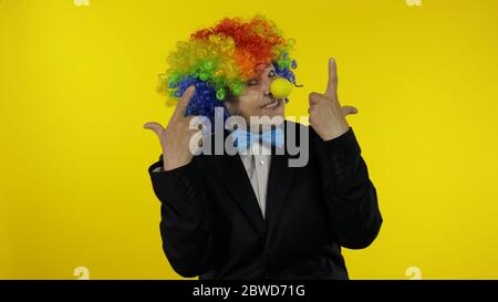 Senior kaukasischen weiblichen Clown in bunten Perücke und blaue Fliege machen dumme Gesichter. Ältere Frau, die Spaß hat, lächelt, tanzt, die Kamera anschaut. Ausdrücke. Kopierbereich. Halloween. Gelber Hintergrund Stockfoto