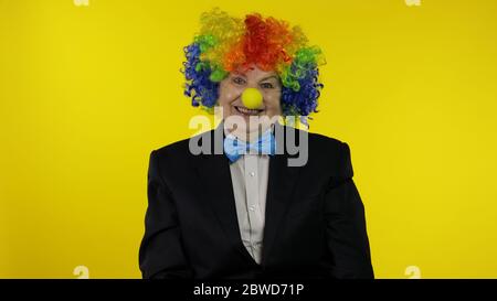 Senior kaukasischen weiblichen Clown in bunten Perücke und blaue Fliege machen dumme Gesichter, Narr um. Ältere Frau, die Spaß hat, lächelt, schaut Kamera an. Kopierbereich. Halloween. Gelber Hintergrund Stockfoto