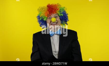 Senior kaukasischen weiblichen Clown in bunten Perücke und blaue Fliege machen dumme Gesichter, Narr um. Ältere Frau, die Spaß hat, lächelt, schaut Kamera an. Kopierbereich. Halloween. Gelber Hintergrund Stockfoto