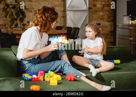 Lockige Mutter hält Spielzeugpistole und sieht beleidigte Tochter mit verschränkten Armen Stockfoto