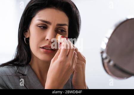 Selektiver Fokus der traurigen Frau mit Blutergüssen auf Gesicht halten Make-up Schwamm während der Anwendung Make-up Stiftung in der Nähe Spiegel, häusliche Gewalt Konzept Stockfoto