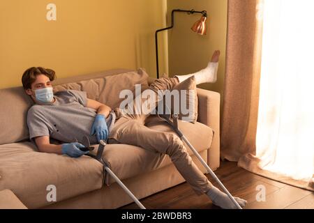 Behinderter Mann in medizinischer Maske und Pflaster Verband am Bein Film in der Nähe Krücken zu Hause Stockfoto