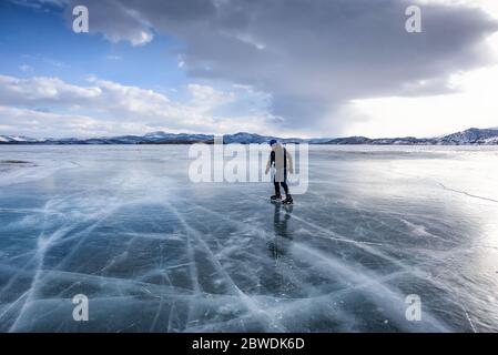 Baikalsee, Russland - 13. März 2020: Mann Eislaufen auf dem gefrorenen Baikalsee mit schneebedeckten Bergen im Hintergrund Stockfoto